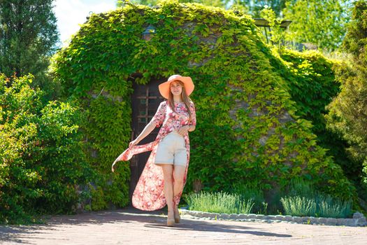 A charming girl in a light summer sundress and a pareo hat is walking in a green park. Enjoys warm sunny summer days.