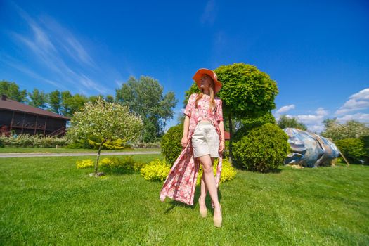 A charming girl in a light summer sundress and a pareo hat is walking in a green park. Enjoys warm sunny summer days.