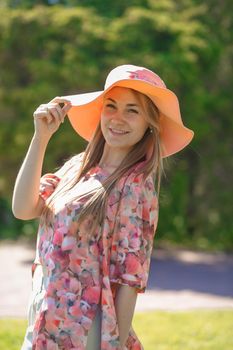 A charming girl in a light summer sundress and a pareo hat is walking in a green park. Enjoys warm sunny summer days.