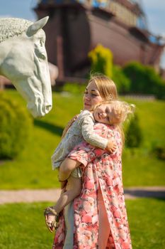 A charming girl in a light summer sundress walks in a green park with her little daughter, holding her in her arms. Enjoys warm sunny summer days.