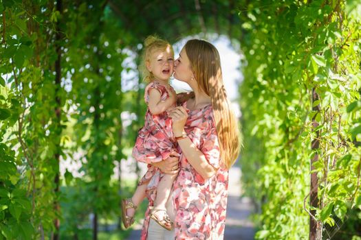 A charming girl in a light summer sundress walks in a green park with her little daughter, holding her in her arms. Enjoys warm sunny summer days.