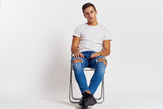 Man on a chair indoors torn jeans white t-shirt handsome face model light background. High quality photo