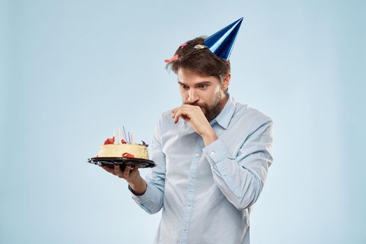 Bearded man with a plate of cake on a blue background and a birthday party hat on his head. High quality photo