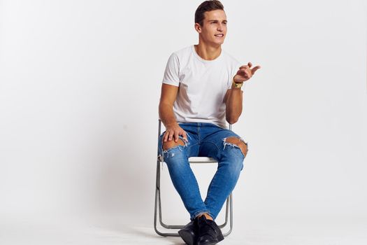 Man on a chair indoors torn jeans white t-shirt handsome face model light background. High quality photo