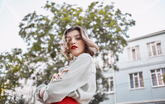 woman with red lips in a fashionable shirt near the building and green trees spring nature. High quality photo