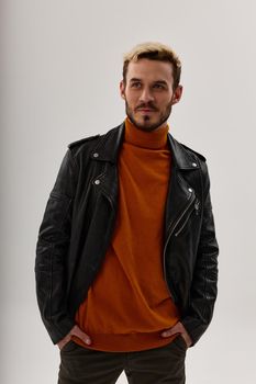 a man in an orange sweater and a leather jacket holds his hands in his pockets on a light background. High quality photo