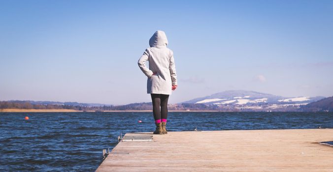 Young girl with grey coat is standing on a footbridge and enjoys the view over the lake, winter time