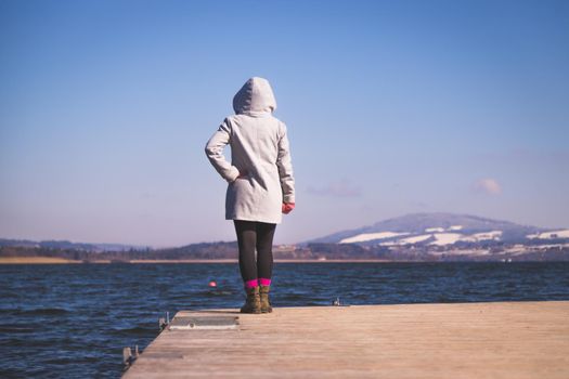 Young girl with grey coat is standing on a footbridge and enjoys the view over the lake, winter time