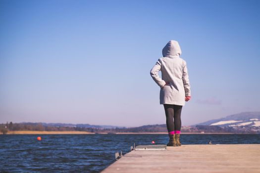 Young girl with grey coat is standing on a footbridge and enjoys the view over the lake, winter time