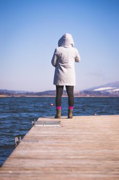 Young girl with grey coat is standing on a footbridge and enjoys the view over the lake, winter time