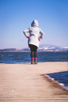 Young girl with grey coat is standing on a footbridge and enjoys the view over the lake, winter time