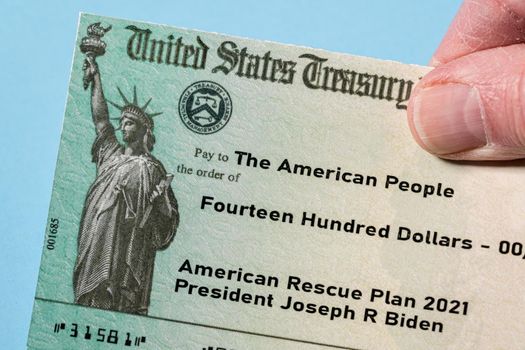 Hand holding a US Treasury illustrative check to illustrate American Rescue Plan Act of 2021 payment on blue background
