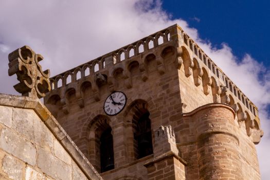 A cross on the top of a stone wall in front of a clock tower against a blue sky on a sunny day