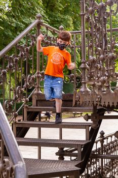 A cute redhead boy wearing a face mask and an orange t-shirt walking the stairs of a metal bandstand on a sunny day