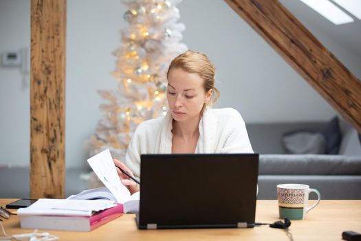 Young businesswoman wearing cosy warm bathrobe working remotly from home in winter Christmas time during 2020 corona virus pandemic. Work from home, selter in place, concept.