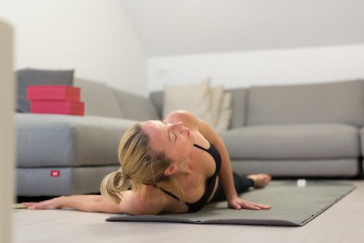 Beautiful blonde woman doing home workout indoors. Woman practice yoga at home. Fit girl using workout tutorials for healthy active lifestyle. Woman using quarantine for home workouts. Stretching