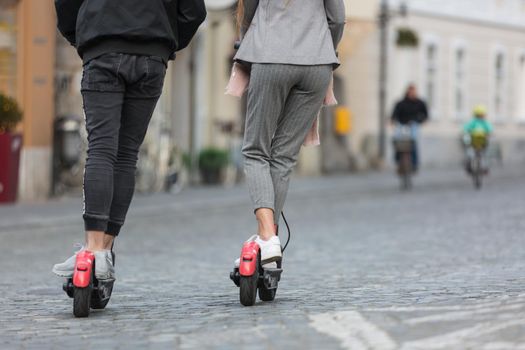 Rear view of unrecognizable trendy fashinable teenagers riding public rental electric scooters in urban city environment. New eco-friendly modern public city transport in Ljubljana, Slovenia.