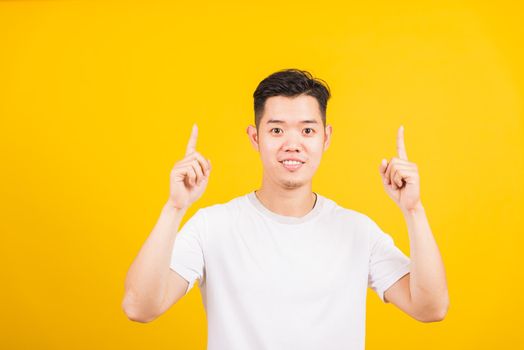 Portrait happy Asian handsome young man smiling standing wearing white t-shirt pointing finger to above UP side he looking to camera, studio shot isolated yellow background