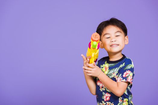 Happy Asian little boy holding plastic water gun, Thai kid funny hold toy water pistol and smiling, studio shot isolated on purple background, Thailand Songkran festival day national culture party