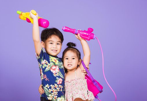 Two Happy Asian little boy and girl holding plastic water gun, Thai children funny hold toy water pistol and smile, studio shot isolated on purple background, Thailand Songkran festival day culture.