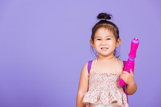 Happy Asian little girl holding plastic water gun, Thai child funny hold toy water pistol and smile, studio shot isolated on purple background, Thailand Songkran festival day national culture concept