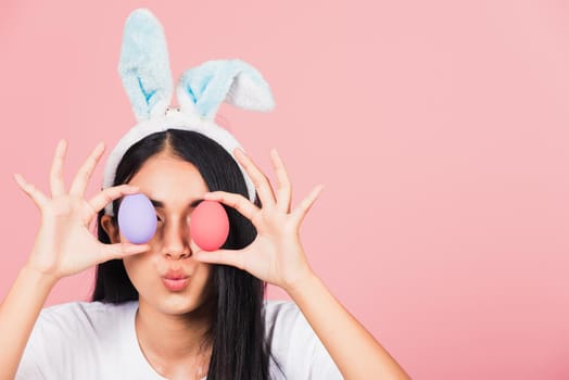 Happy beautiful young woman smiling wearing rabbit ears holding colorful Easter eggs front eyes, Thai female with bunny ear hold easter egg covering eye, studio shot isolated on pink background