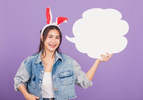 
Happy Easter Day. Beautiful young woman smiling excited wearing rabbit ears and denims holding empty speech bubble, Portrait female looking at camera, studio shot isolated on purple background