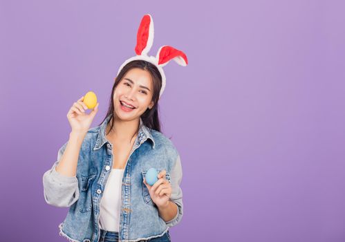 Happy Easter concept. Beautiful young woman smiling wearing rabbit ears and denims hold colorful Easter eggs gift on hands, Portrait female looking at camera, studio shot isolated on purple background