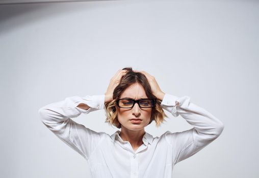 A woman in glasses and in a white shirt touches her head with her hands on a light background. High quality photo
