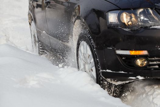 Black vehicle makes its way through the deep snow rut skidding, car's wheels spin and spew up pieces of snow it attempts to gain traction on the slippery road.
