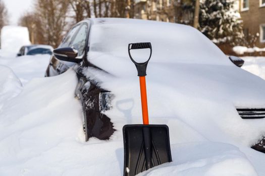 plastic snow shovel in front of snow-covered car at sunny winter morning, close-up with selective focus