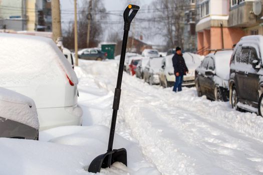 big black plastic snow shovel near snow-covered cars at sunny winter morning while people cleaning their cars out of snow