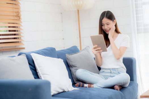 Young attractive asian woman resting using browsing tablet computer on sofa at home, happy girl sitting on couch relax reading digital gadget with excited, communication and lifestyle concept.