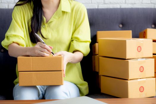 Closeup hand of young asian woman writing address on parcel box at home for delivery order to customer, shipping and logistic, merchant online and seller, business owner or SME, online shopping.