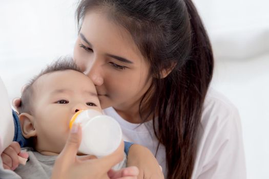 Young asian mother embracing and feeding little baby girl with bottle of milk at home, newborn innocence drinking with mom satisfied, relationship and bonding of mum and child, family concept.