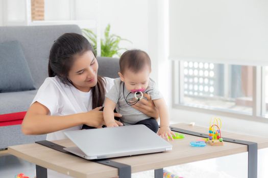 Young asian mother working on laptop computer while care daughter girl at home, mom and baby girl sitting using notebook video call, parent and bonding, indoors, family and business concept.