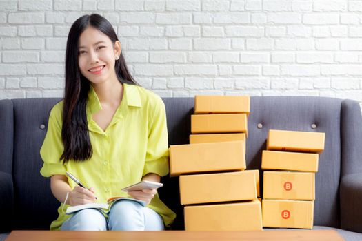 Young asian woman is entrepreneur looking phone and writing order of customer with online on notebook at home, online shopping, logistic and delivery, female is seller working on sofa, business SME.