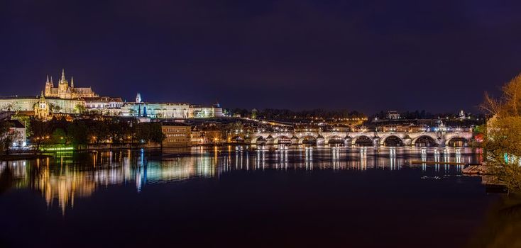 Charles Bridge in Prague in the evening with colorful lights from lanterns. In the river the reflection of the evening illuminations. Panorama