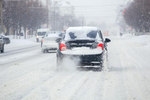 Road ice pieces flow from wheels of snow covered black car moving fast in daylight city with selective focus. Vehicle moving on snowy highway after heavy winter snowfall.