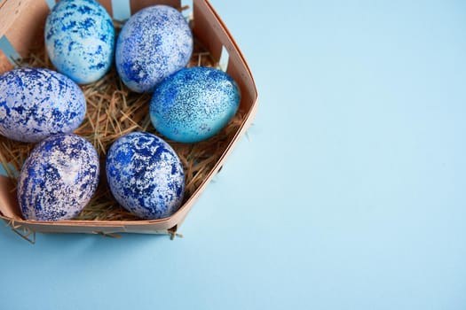 Blue chicken eggs lie in round wooden basket which stands on a blue paper background. Easter background. Seasonal holiday flat lay with free space for text. Classic blue color of the year 2020
