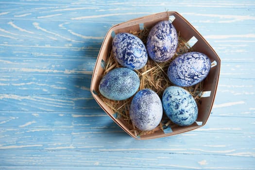 Blue chicken eggs lie in round wooden basket which stands on a blue wooden table. Easter background. Seasonal holiday flat lay with free space for text. Classic blue color of the year 2020