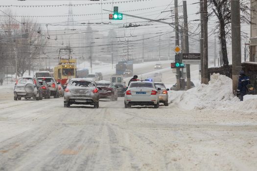 Tula, Russia - February 13, 2020: City vehicles stopped at crossroads in front of traffic lights with police car during heavy snowfall at daytime.