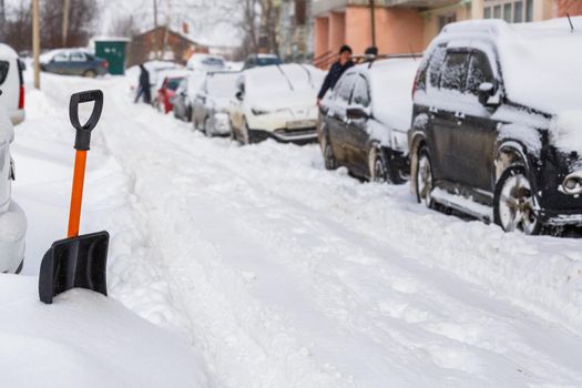 small plastic snow shovel near snow-covered cars at sunny winter morning while people cleaning their cars out of snow