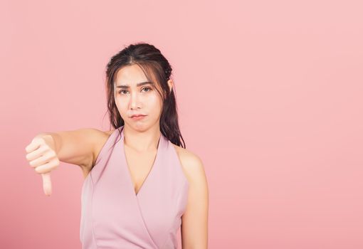 Portrait Asian beautiful young woman unhappy, negative gesture showing finger thumbs down or dislike sign, studio shot isolated on pink background, Thai female rejection unlike with copy space