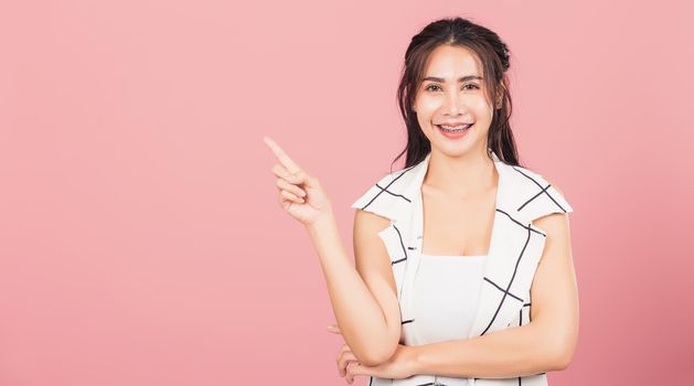 Happy Asian portrait beautiful cute young woman standing smiling indicate finger empty space looking to camera, studio shot isolated on pink background, Thai female pointing index out with copy space