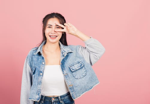 Asian happy portrait beautiful cute young woman wear denim smile standing showing finger making v-sign victory symbol near eye looking to camera studio shot isolated on pink background with copy space