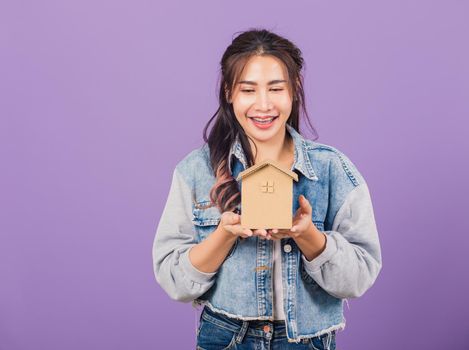 Happy Asian portrait beautiful cute young woman wear denim excited smiling holding house model on hand, studio shot isolated on purple background, broker female hold home real estate insurance