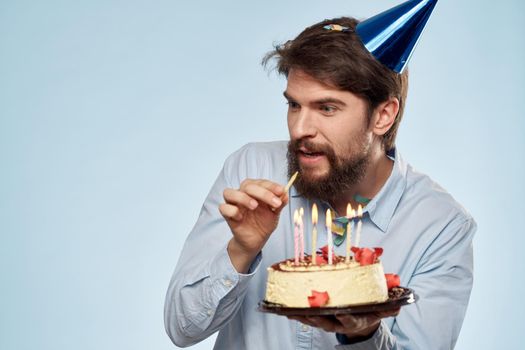 Bearded man with a plate of cake on a blue background and a birthday party hat on his head. High quality photo
