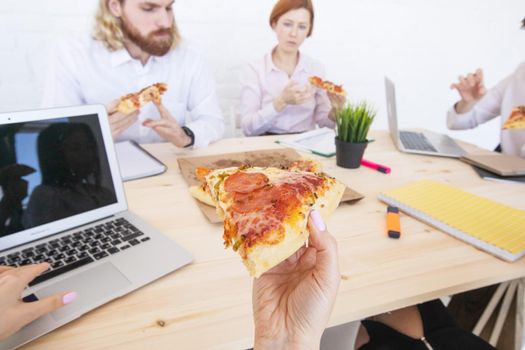 Group of diverse business people eating pizza at break while working together in an office sitting around desk