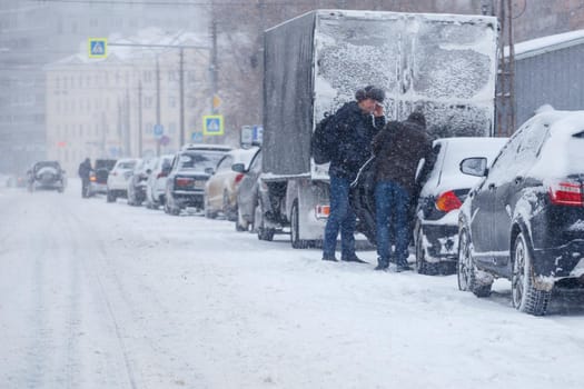 Tula, Russia - February 13, 2020: Two men standin near car parked on side of city road during heavy snowstorm at day light.